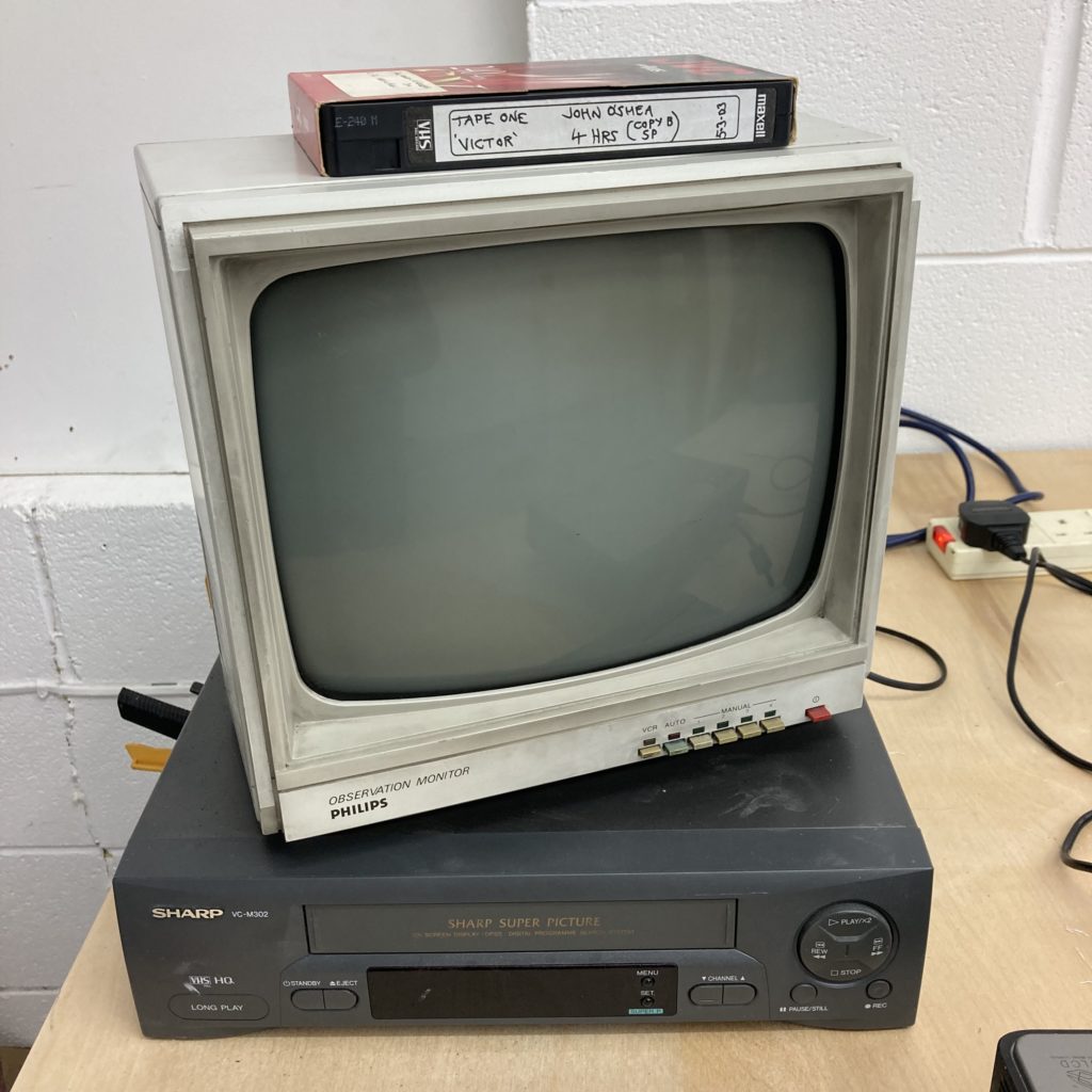 a VHS tape on top of a 1990's CCTV black and white crt monitor on top of a grey Sharp VHS player.